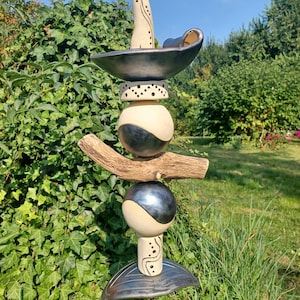 Può includere: Una scultura da giardino in ceramica con un design fantasioso. Presenta un pezzo alto e curvo con vortici neri e bianchi, una ciotola nera e una serie di sfere nere e bianche. La scultura è montata su un ramo di legno e un paletto di metallo.