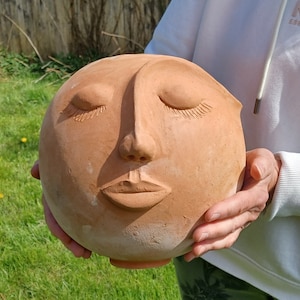Könnte beinhalten: Eine Terrakotta-Skulptur eines Gesichts mit geschlossenen Augen und leicht geöffnetem Mund. Die Skulptur wird in der Hand gehalten und scheint aus Ton zu sein.