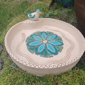 Puede incluir: Un bebedero para pájaros de cerámica blanca con un diseño floral azul y verde. Una pequeña figurita de pájaro azul se sienta en el borde del bebedero.