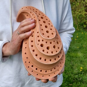 Könnte beinhalten: Eine terrakottafarbene, abstrakte Skulptur mit einer geschichteten, organischen Form. Die Skulptur weist zahlreiche kreisförmige Perforationen auf. Das Stück wird vor einem weißen Kapuzenpullover und einem grünen Hintergrund gehalten.