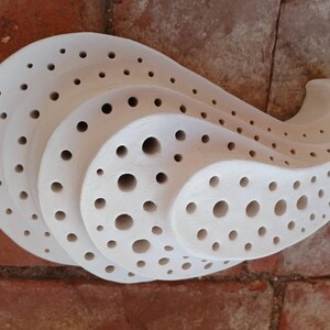 May include: White ceramic wall sculpture with a unique, organic shape and a pattern of small, round holes. The sculpture is designed to resemble a shell or a wave.