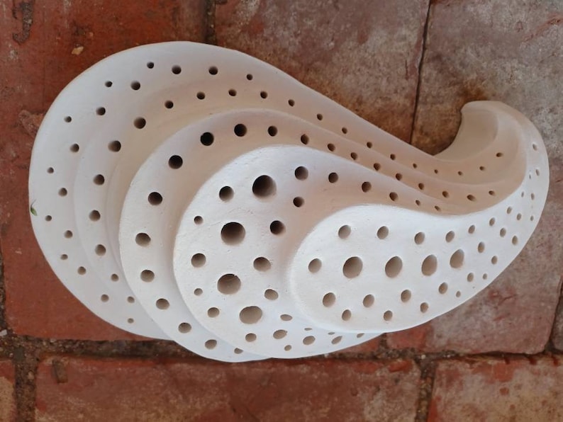 May include: White ceramic wall sculpture with a unique, organic shape and a pattern of small, round holes. The sculpture is designed to resemble a shell or a wave.