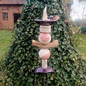 Può includere: Una scultura da giardino artigianale e stravagante. Presenta un design impilato con elementi in ceramica nei toni del viola, rosa e bianco, accentuati da un ramo di legno. La scultura è ambientata su uno sfondo di verde lussureggiante.