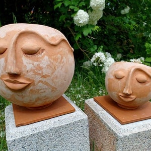 Könnte beinhalten: Zwei Terrakotta-Pflanztopf in Form von Gesichtern mit geschlossenen Augen und lächelnden Mündern. Der größere Topf steht auf einem quadratischen Sockel, der kleinere Topf auf einem rechteckigen Sockel.