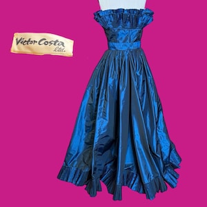 May include: A vintage, floor-length, navy blue gown with a ruffled neckline and hem. The dress features a fitted bodice and a full skirt, cinched at the waist with a matching belt. The label "Victor Costa ltd." is visible in the upper left corner.