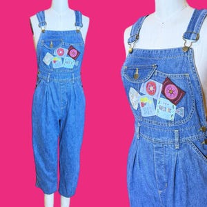 May include: Light blue denim overalls with decorative patches on the bib pocket. The overalls have adjustable straps, side buttons, and a relaxed fit. The patches feature various designs, including stars and geometric shapes. The text "GUESS INC" is visible on one of the patches.
