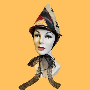 May include: A unique, pointed hat with a patchwork design in shades of beige, red, black, and yellow. The hat is adorned with black star-shaped accents and a black lace trim. A black lace bow is tied around the neck of a mannequin head.