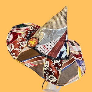 May include: A patchwork hat with a variety of fabrics in shades of brown, red, and cream. The hat features a decorative pin with a gold emblem and black lace trim. A gold chain is attached to the brim.