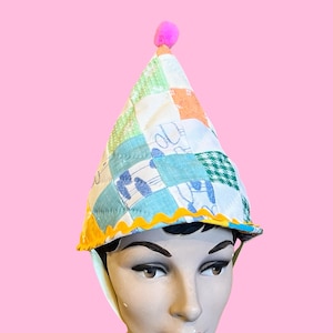 May include: A patchwork party hat with a pink pom-pom on top. The hat is made of various fabric squares in pastel colors, including blue, green, orange, and white. A yellow, scalloped trim lines the bottom edge.