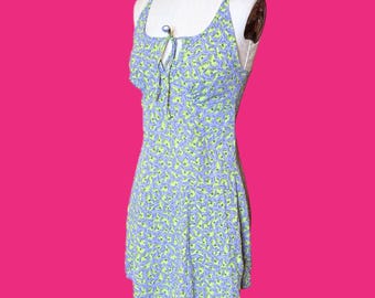 May include: A sleeveless, mini dress with a scoop neckline and a tie detail. The dress features a blue and green floral pattern. The dress is displayed on a mannequin against a pink background.