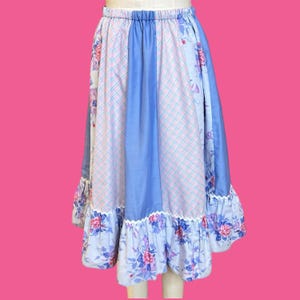 May include: A vintage skirt with a patchwork design. The skirt features panels of light blue, pink plaid, and floral fabric. A ruffled hem with floral print and white lace trim completes the look. The skirt has an elastic waistband.