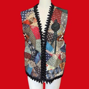 May include: A sleeveless vest constructed from a patchwork of various patterned fabrics in shades of red, blue, brown, and cream. The garment features a black braided trim along the front opening and hem, and a black fabric rose pinned to the chest.