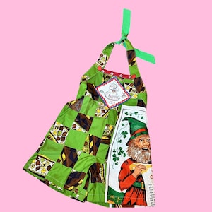 May include: Green and brown patchwork overall dress with a red trim and a small fabric applique of a girl. The dress has a large fabric applique of a leprechaun holding a shamrock and a sign that says "Just a glass of Irish Coffee. Add a little coffee and cream. Enjoy!"