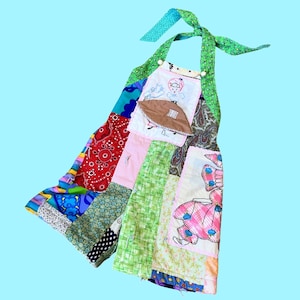 May include: A patchwork quilt apron with a green, red, blue, and pink color scheme. The apron has a halter neck strap and a pocket with an embroidered image of a girl.