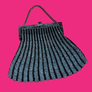 May include: A vintage black and silver beaded handbag with a chain handle. The purse features vertical stripes of black and silver beads, creating a textured, fan-shaped design. The clasp is a decorative silver metal. The background is a bright pink.