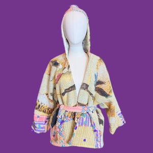 May include: A hooded, open-front jacket with a patchwork design. The jacket features a cream-colored base with colorful patterns, a pink belt, and patterned cuffs. The jacket is displayed on a mannequin against a purple background.