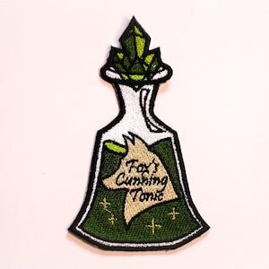 Puede incluir: Parche bordado con un diseño de botella de poción. La botella contiene líquido verde y una cabeza de zorro con el texto "Fox's Cunning Tonic". La parte superior de la botella presenta formas de cristal verde. El parche tiene un borde negro.