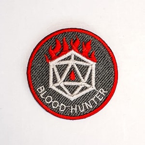 Puede incluir: Parche redondo bordado con fondo negro y borde rojo. El parche presenta una forma geométrica blanca con una gota roja, rodeada de llamas rojas. Las palabras "BLOOD HUNTER" están bordadas en blanco.