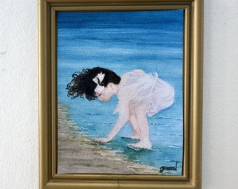 May include: A framed painting of a young girl in a white dress with a black bow in her hair, standing in the water at the edge of a beach. The water is blue and the sand is light brown.