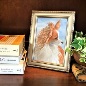 May include: A framed watercolor painting of a long-haired, brown and white papillon dog with its head turned to the side. The dog is looking to the right of the image. The painting is set on a wooden shelf with books and a potted plant.