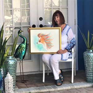 May include: A woman holds a framed watercolor painting of a dog with colorful fur. The painting is in a gold frame and the dog is looking to the left. The woman is sitting on a white chair in front of a door with a window. There are two green plants in pots on either side of the door.