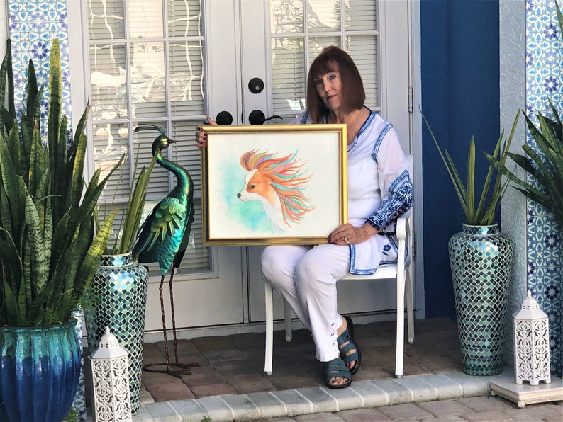 May include: A woman holds a framed watercolor painting of a dog with colorful fur. The painting is in a gold frame and the dog is looking to the left. The woman is sitting on a white chair in front of a door with a window. There are two green plants in pots on either side of the door.