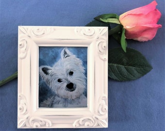 Könnte beinhalten: Ein weiß gerahmtes Gemälde eines weißen Hundes vor blauem Hintergrund. Eine rosa Rose mit grünen Blättern liegt neben dem Rahmen.