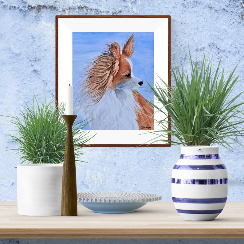 May include: A watercolor painting of a Papillon dog with brown and white fur, looking to the right. The painting is framed in a brown wood frame and is hanging on a blue wall.