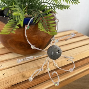 May include: A decorative beaded ornament with a dark gray sphere center and radiating clear beaded arms with iridescent accents. The ornament is placed on a wooden slatted surface near a wooden bowl with a green plant.