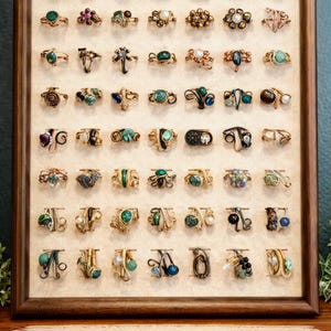 May include: A display of handcrafted wire-wrapped rings, each unique in design and gemstone. The rings are arranged in a wooden framed display, showcasing a variety of colors and styles, with the text "HANDCRAFTED WIRE-WRAPPED RINGS" at the bottom.
