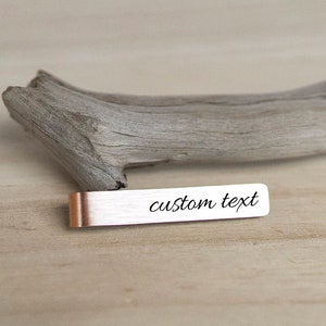 May include: A silver tie bar with a custom text inscription on a wooden surface.