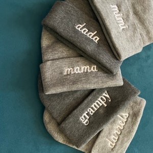 May include: A stack of gray knit beanies with white embroidered text. The beanies display the words "dada", "mama", "grampy", "darrels", and "mimi". The hats are arranged on a teal blue surface.