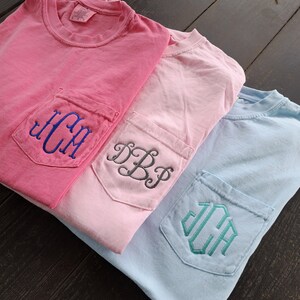 May include: Three t-shirts with different colored pockets and embroidered monograms. The top shirt is pink with a blue monogram "JCM". The middle shirt is light pink with a gray monogram "DBP". The bottom shirt is light blue with a teal monogram "JCA".