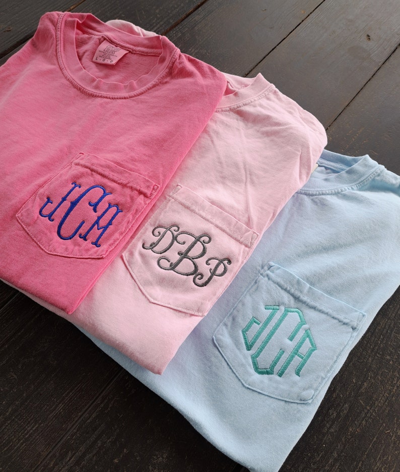 May include: Three t-shirts with different colored pockets and embroidered monograms. The top shirt is pink with a blue monogram "JCM". The middle shirt is light pink with a gray monogram "DBP". The bottom shirt is light blue with a teal monogram "JCA".