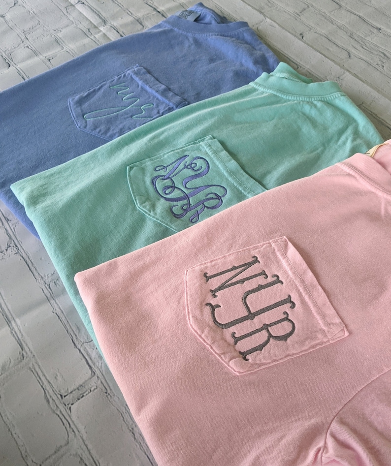 May include: Three t-shirts with different colors and embroidered monograms on the pocket. The shirts are stacked on top of each other, with the blue shirt on top, followed by the green shirt, and then the pink shirt.
