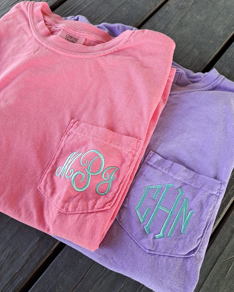 May include: Two t-shirts with embroidered monograms on the front pockets. The top shirt is pink with the monogram "M.P.G." and the bottom shirt is purple with the monogram "C.H.N."