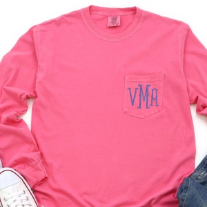 May include: Pink long-sleeve t-shirt with a pocket on the left side. The pocket has a blue monogram that reads "VMM".
