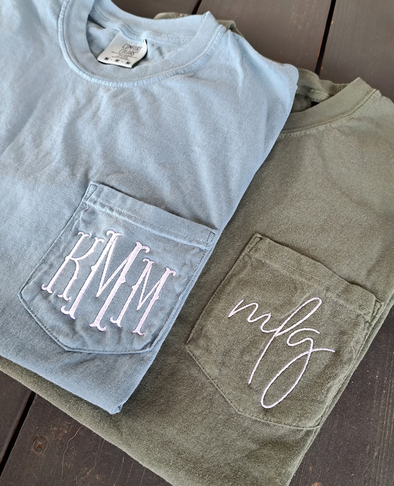 May include: Two t-shirts with pockets. The top shirt is light blue with a white monogram "KMM" embroidered on the pocket. The bottom shirt is olive green with a white cursive monogram "mfg" embroidered on the pocket.