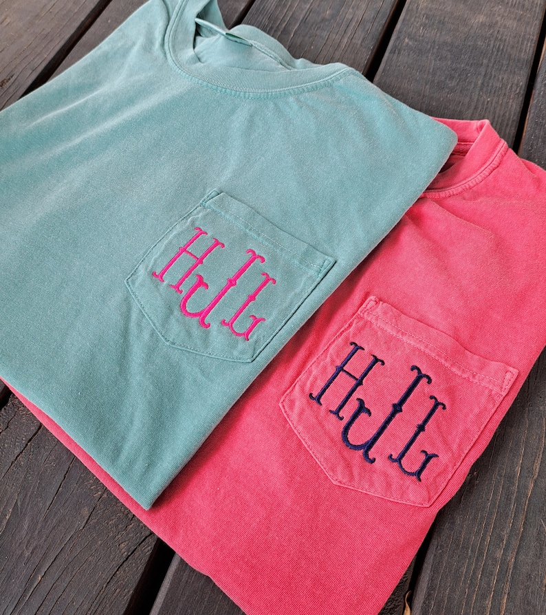 May include: Two t-shirts with a pocket on the front. The top shirt is teal with a pink monogram that says "HLL". The bottom shirt is red with a navy blue monogram that says "HLL".