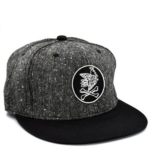 Pode incluir: Um boné de beisebol de tweed preto e cinza com aba preta. O boné tem um remendo bordado branco de um pássaro com halo e ossos cruzados.