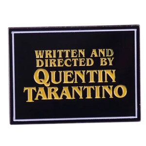 May include: Black and white enamel pin with a white border. The pin features gold text that reads "Written and Directed by Quentin Tarantino".