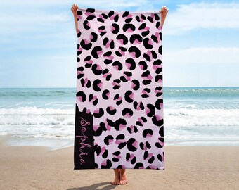 May include: A beach towel with a pink leopard print design. The towel features black and pink spots on a light pink background. A black vertical stripe on the side displays the name "Sophia" in cursive.