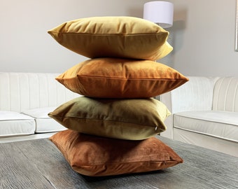 May include: A stack of four square velvet throw pillows in shades of gold, orange, and brown. The pillows are arranged on top of each other, with the bottom pillow resting on a wooden surface. The pillows have a soft, plush appearance.
