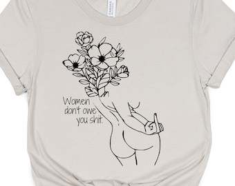 Peut inclure: T-shirt beige avec un dessin au trait noir d'une personne tenant des fleurs et faisant un doigt d'honneur. Le texte "Women don't owe you shit" est imprimé sur le t-shirt.