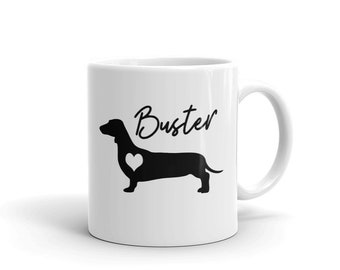 May include: White ceramic mug with a black silhouette of a dachshund with a heart in its chest. The text "Buster" is written in black cursive above the dog.