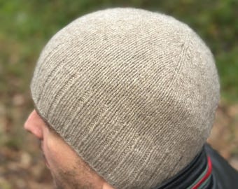 May include: A light brown knit beanie hat. The hat is made of a ribbed knit material and has a rounded crown. The hat is worn by a person wearing a black leather jacket with a red and green stripe.