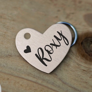 May include: A silver heart-shaped pet tag with the name "Roxy" engraved in black lettering. A small black heart is above the name.