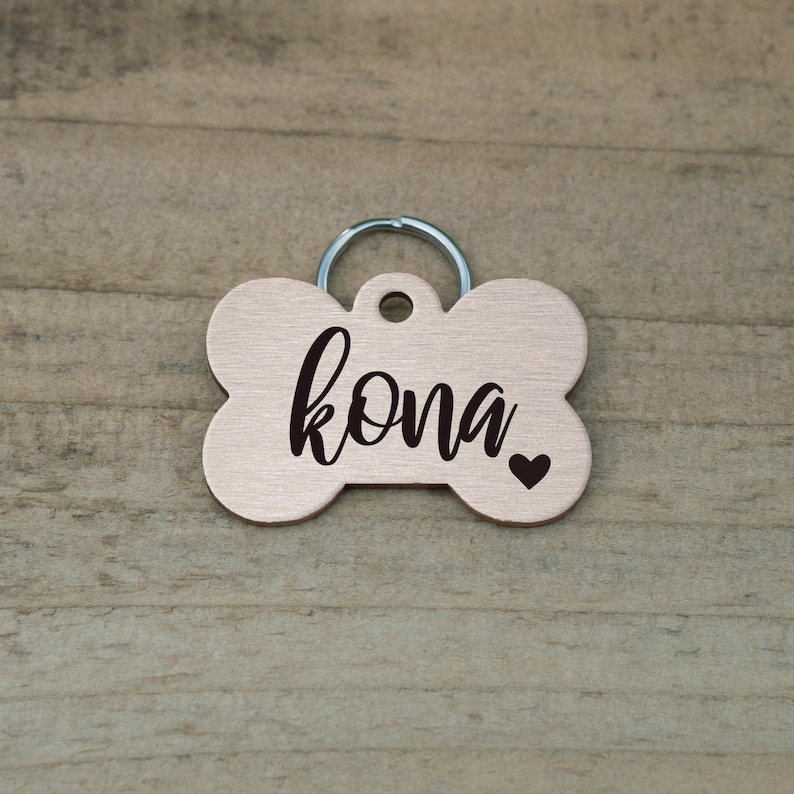 May include: A rose gold bone-shaped dog tag with the name "Kona" engraved in black script and a small black heart.