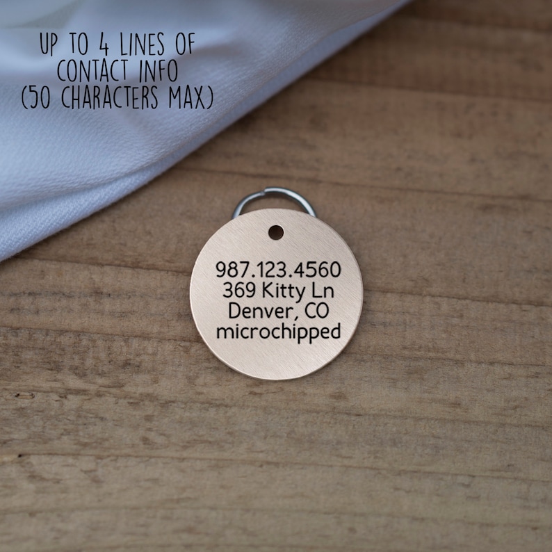 Pode incluir: Uma etiqueta redonda para animais de estima&ccedil;&atilde;o de cor cobre com texto gravado. O texto diz: "987.123.4560 369 Kitty Ln Denver, CO microchipped".