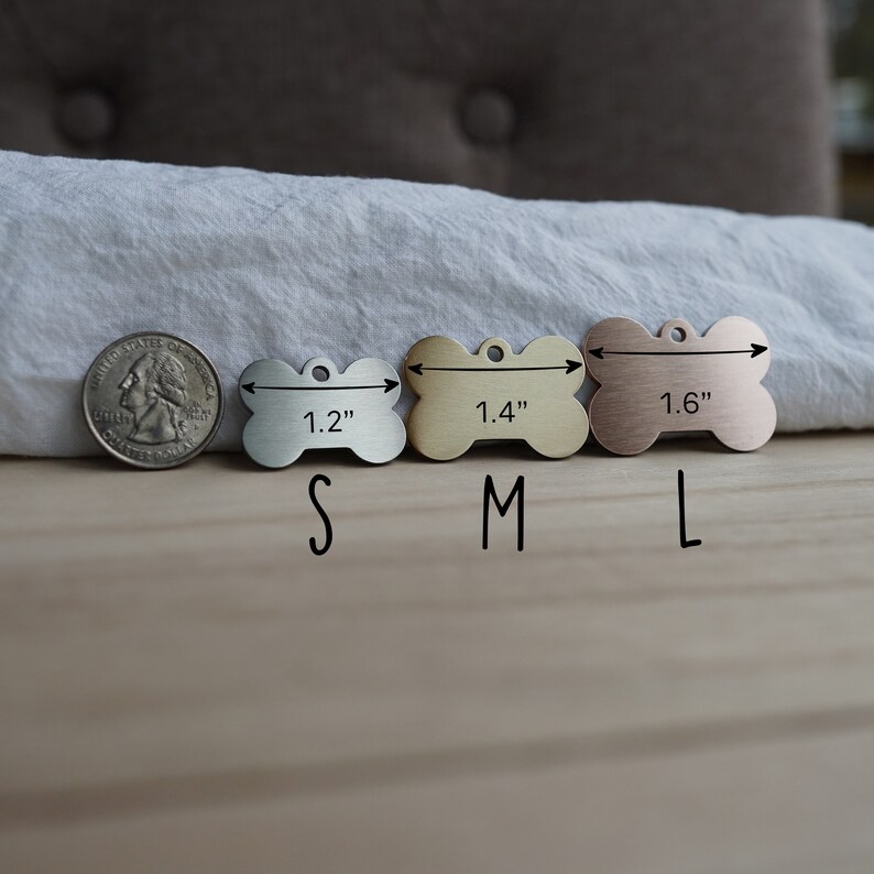 May include: Three silver dog bone-shaped tags with measurements of 1.2 inches, 1.4 inches, and 1.6 inches. The tags are arranged from smallest to largest. The tags are on a wooden surface.